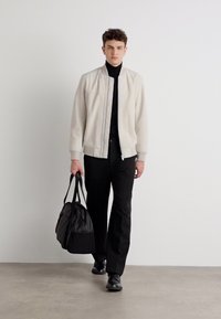 Heled hall bomber-jakk, must turtleneck, must püksid ja mustad kingad. Kätel must kott, seismine neutraalses taustas.