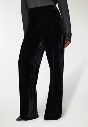 Donna in pantaloni neri a gamba larga di velluto e una maglietta a maniche lunghe grigia scura, in piedi con una mano parzialmente visibile al fianco.