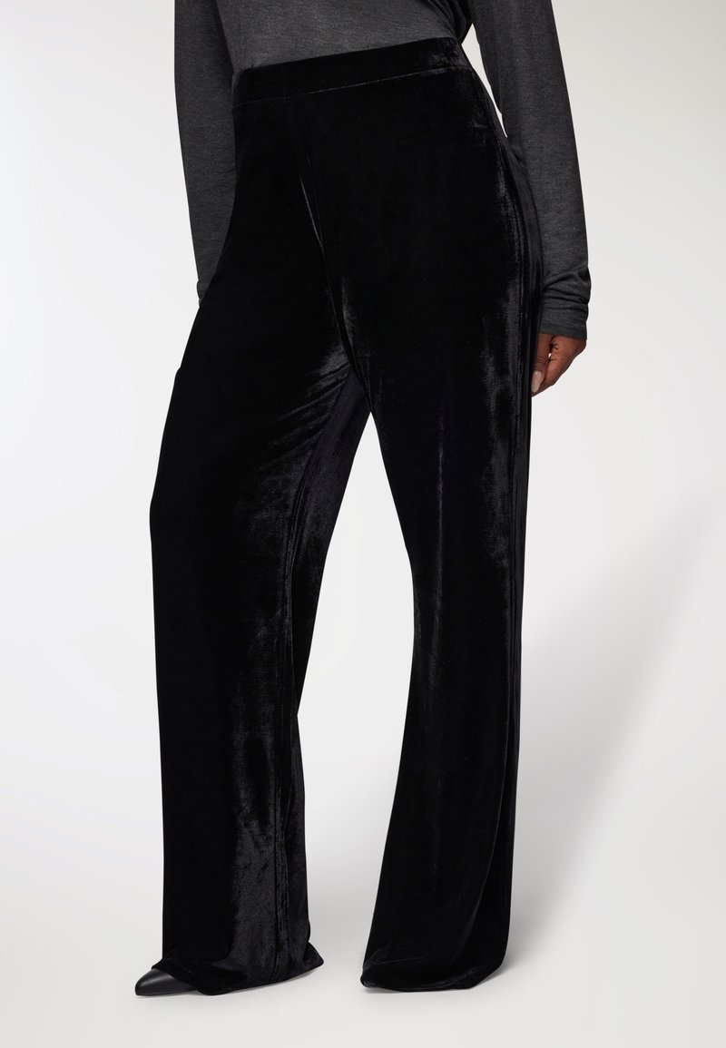 Kobieta w czarnych, szerokich, aksamitnych spodniach oraz długim, ciemnoszarym topie, stojąca z jedną ręką częściowo widoczną przy boku.