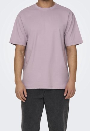 Person iført en ensfarvet lys lilla T-shirt med korte ærmer og mørkegrå bukser, vist fra nakken til midt på låret mod en hvid baggrund.