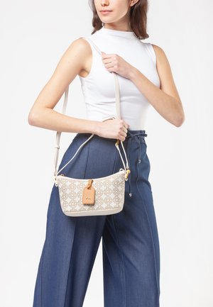 Mujer con top blanco sin mangas y pantalones azules sostiene un bolso bandolera beige con patrón geométrico y etiqueta de cuero marrón claro.