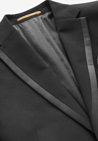 Chaqueta de traje negra con solapas de satén, que presenta un forro interior gris con patrón, textura suave y elementos de diseño estructurados.