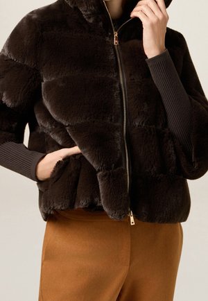 Personne portant une veste zippée en peluche marron foncé avec des poignets côtelés et un pantalon marron clair, une main dans la poche et l'autre près du visage.
