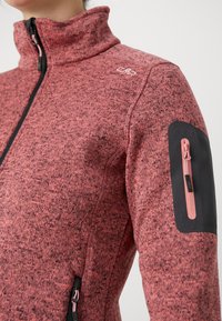 Tekstuurilla varustettu korkeakauluksellinen fleece-takki, jossa on raitakuoseja pinkissä sävyssä. Takissa on musta käsitasku, jossa on vaaleanpunainen vetoketju, sekä sivuilla vetoketjulliset taskut.