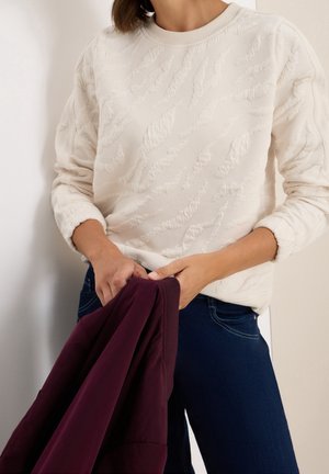 Personne portant un pull texturé blanc cassé et un pantalon bleu foncé tenant un vêtement plié violet foncé sur un fond clair.