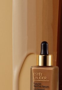 Estee Lauder Futurist SkinTint Serum i en frostet glassflaske med gullfarger, pipette-topp og en varm brun nyanse. Glatt tekstur.