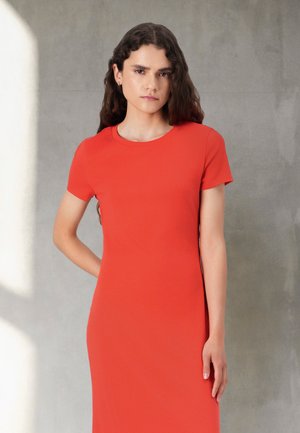 Woman with long curly hair wearing a fitted short-sleeve red dress, standing against a plain light gray wall with one hand behind her back.