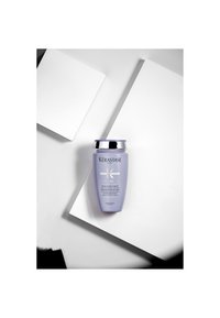 Flasche des lila Shampoos Kérastase Blond Absolu Bain Ultra-Violet, platziert auf schrägen weißen geometrischen Flächen.