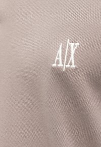 Svetlo béžová textúrovaná látka s vyšívaným bielym logom "A/X". Povrch má jemný vzor tkania, ktorý zdôrazňuje kvalitu materiálu.