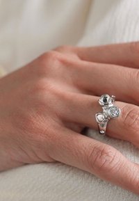 Inel din argint sterling, cu trei pietre distincte: o piatră rotundă transparentă, o inimă argintie și o piatră cu design floral, evidențiind detalii complicate.