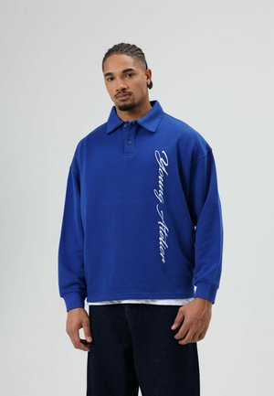Blauw oversized poloshirt met lange mouwen, kraag en wit bedrukt tekst aan de zijkant. Gemaakt van zachte stof met een ontspannen pasvorm.
