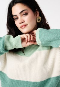 Pull en maille épaisse avec des rayures horizontales vert menthe et crème. Gros plan sur le tissu texturé et les poignets côtelés. Des boucles d'oreilles audacieuses accentuent les détails.