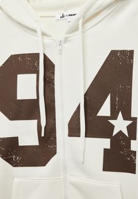Weißer Zip-Hoodie mit großem braunem "94" und Stern-Grafik. Mit Kordeln, weichem Material und Vordertasche für praktischen Nutzen.