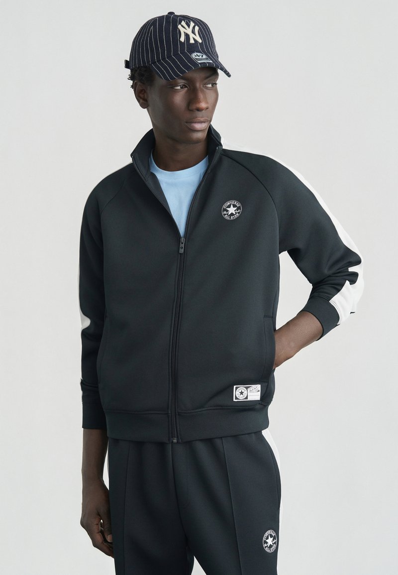 Μαύρο φούτερ με φερμουάρ και λευκές ρίγες στους ώμους, διακοσμημένο με στρογγυλή ετικέτα λογότυπου. Συνδυασμένο με μαύρη φόρμα jogger, φορεμένο με ριγωτό καπελάκι.