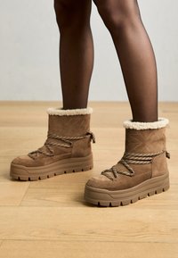 Bottes chelsea en daim marron avec doublure en fourrure, semelles en caoutchouc épaisses et lacets tressés ; surface texturée avec un motif matelassé sur la tige.