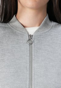 Nærbilde av en persons nakke og bryst som har på seg en grå hettegenser med glidelås, som har en rund glider og en ribbestrikket krage under.