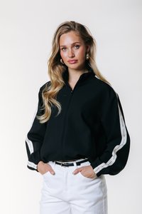 Chaqueta negra con cremallera y cuello alto, rayas laterales blancas y puños elásticos, combinada con pantalones blancos y un cinturón negro. Texture suave.
