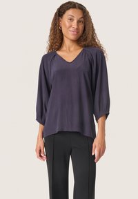 Blusa blu navy a V con maniche a tre quarti, in tessuto leggero e dettagli arricciati sulle spalle. Abbinata a pantaloni neri.