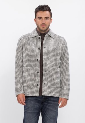 Man draagt een grijs-wit patroon jasje met knopen over een donker shirt en blauwe spijkerbroek, staand tegen een effen witte achtergrond.