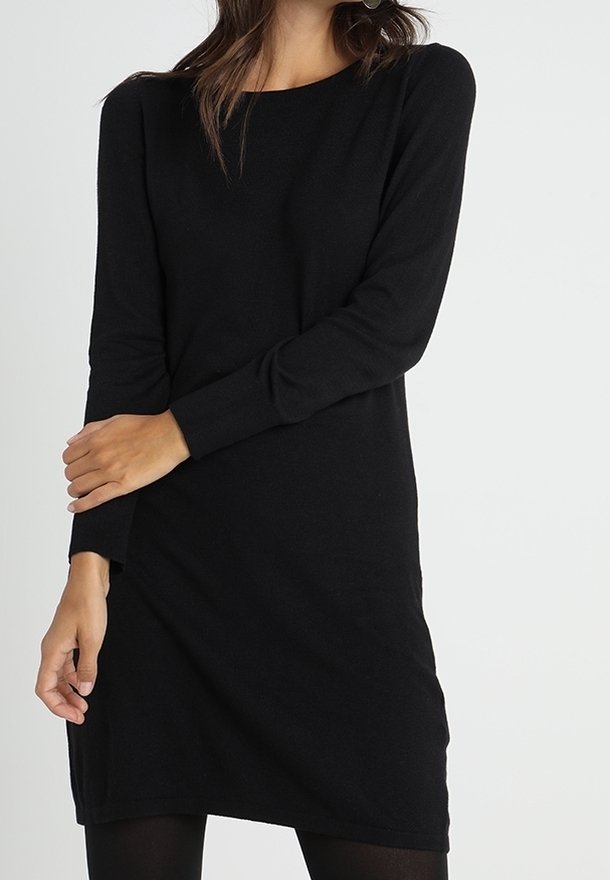 Femme portant une robe noire simple à manches longues, debout avec un bras croisé sur l'autre, sur un fond blanc.