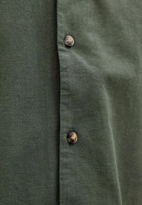 Chemise en lin vert avec un tissu texturé, dotée d'une rangée de boutons en contraste brun et d'accents foncés le long de la patte de boutonnage.