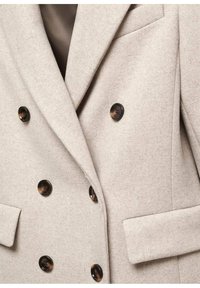 Manteau beige croisé avec six boutons en écaille foncée et poches à rabat sur chaque côté.