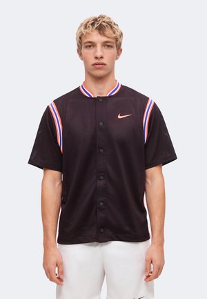 Ung mand iført sort sportstrøje med knapper med rød, hvid og blå kant og Nike-logo, kombineret med hvide shorts.