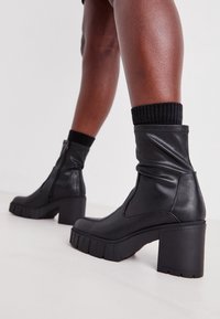 Botas de cuero negro hasta el tobillo con un tacón grueso, que cuentan con una cremallera, un diseño arrugado y una suela de goma gruesa. Puño negro acanalado visible.