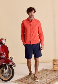 Chemise à manches longues en corail avec des boutons, shorts navy et chaussures lacées beige. Debout à côté d'une trottinette rouge sur un tapis à motifs.
