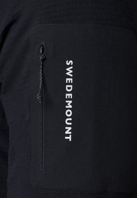 Black fabric panel with vertical white "SWEDEMOUNT" text and a black zipper pocket with a cord pull tab on dark clothing.