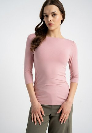 Kobieta o długich brązowych włosach, ubrana w dopasowany różowy top z rękawami do łokcia oraz oliwkowozielone spodnie, stojąca z rękami z przodu.