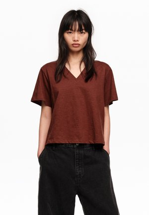 Mujer joven con cabello largo y oscuro, vistiendo una camiseta marrón de manga corta con cuello en V y pantalones negros de cintura alta, con las manos en los bolsillos.