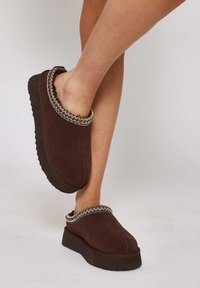 Scarpe slip-on in suede marrone con punta arrotondata, interno morbido e una decorazione in trama intrecciata lungo il colletto. Suola in gomma nera spessa.