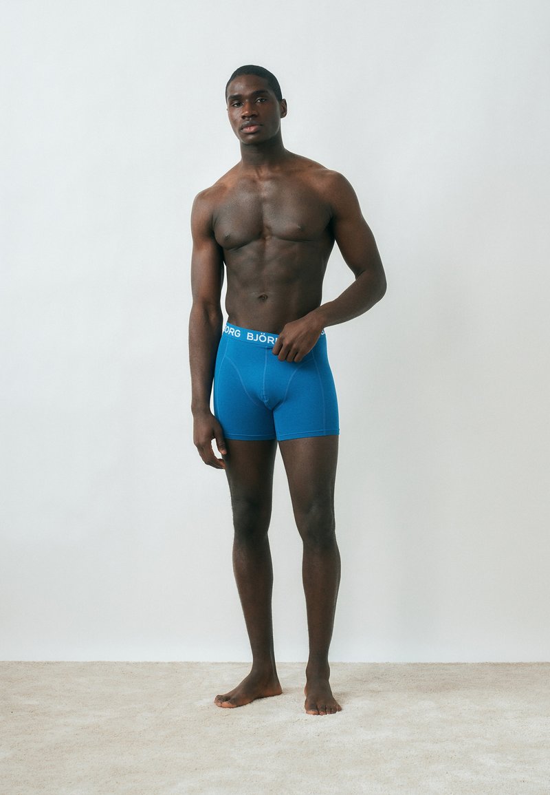 Όρθιος άνδρας χωρίς πουκάμισο, που φοράει μπλε μποξεράκι Björn Borg, στέκεται σε ανοιχτόχρωμο χαλί με φόντο έναν απλό λευκό τοίχο.
