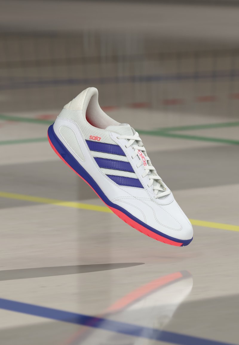 Zapatilla deportiva blanca con rayas moradas, detalles rojos y parte superior texturizada. Tiene punta redonda y suela plana en rosa y azul.