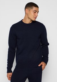 Junger Mann in einem dunkelblauen, gerippten Strickpullover und passender Hose, mit einer Hand in der Tasche, der nach unten schaut und leicht lächelt.