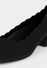 Chaussure pour femme noire à talon bas avec bords festonnés et semelle intérieure texturée sur fond blanc.