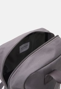 Sac à dos en tissu gris avec une fermeture éclair. L'intérieur présente une doublure lisse, une étiquette cousue à l'intérieur et des poches visibles à l'ouverture.