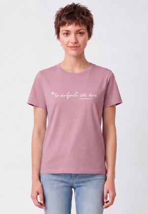 Junge Frau mit kurzen braunen Haaren, die ein mauvefarbenes T-Shirt mit weißem deutschem Text "So einfach ist das" und hellblaue Jeans trägt, steht vor weißem Hintergrund.