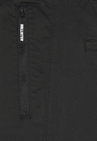 Černé kalhoty vyrobené z texturované látky, které mají zapínací kapsu s nášivkou "HOLLISTER". Obsahují také další detail v podobě boční kapsy.