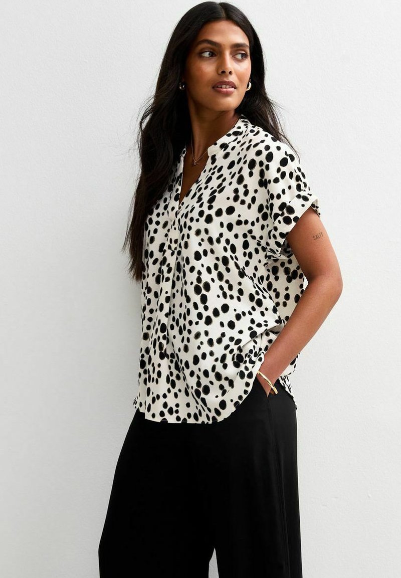 Blusa a maniche corte in tessuto bianco con motivo a pois neri, caratterizzata da scollatura a V e vestibilità rilassata. Abbinata a pantaloni neri.