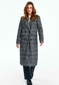 Žena v dlhom sivom károvanom dvojradovom kabáte, čiernych nohaviciach a čiernych topánkach stojaca proti čistému bielemu pozadiu.