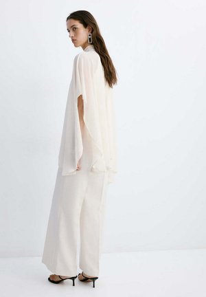 Mujer con cabello largo lleva una blusa tipo capa blanca fluida, pantalones de pierna ancha, pendientes geométricos y sandalias negras de tacón, de pie de lado.