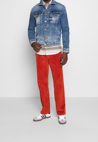 Džínová bunda s lehkým vyblednutím, oranžové manšestrové kalhoty a bílé tenisky s černými pruhy. Oblečení doplňuje vzorovaná košile pod bundou.