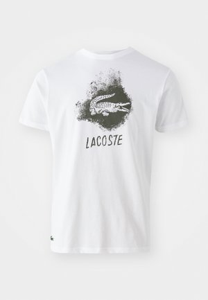 Camiseta de algodón blanca con un gráfico de cocodrilo negro y el texto "LACOSTE", con un logo de acento verde en el dobladillo. Manga corta, cuello redondo.