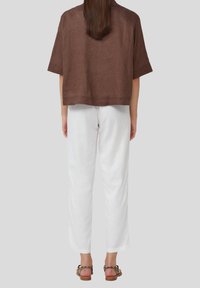 Femme se tenant de dos, portant un haut marron ample, un pantalon blanc droit et des sandales imprimées léopard, le tout sur un fond uni.