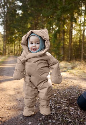 Combinaison de neige rembourrée couleur beige avec une grande capuche, des moufles surdimensionnées et une couche de base à capuche bleue. Déposée sur un chemin forestier avec un doux soleil filtrant à travers les arbres.