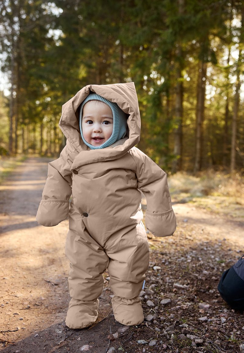 Hnedý nadýchaný snowsuit s veľkou kapucňou, nadrozmernými rukavicami a modrou základnou vrstvou s kapucňou. Umiestnený na lesnej ceste s jemným slnečným svetlom prenikajúcim cez stromy.