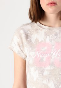 Světle béžové tričko s květinovým vzorem a krátkými rukávy. Vybaveno růžovým textem "New York 88" s vyšitými detaily. Měkká texturální látka.