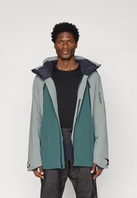 Férfi kéttonusú szürke és türkiz kültéri dzsekiben, fekete pólóval és sötét nadrággal, egy világos, egyszínű háttér előtt áll.